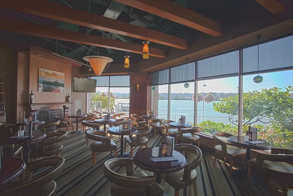 Restaurant interior with dining tables, exposed beams, and panoramic water view.