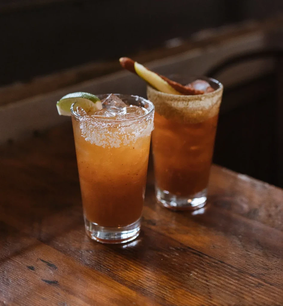 Two amber drinks, garnished rims, lime, and stick on wood.