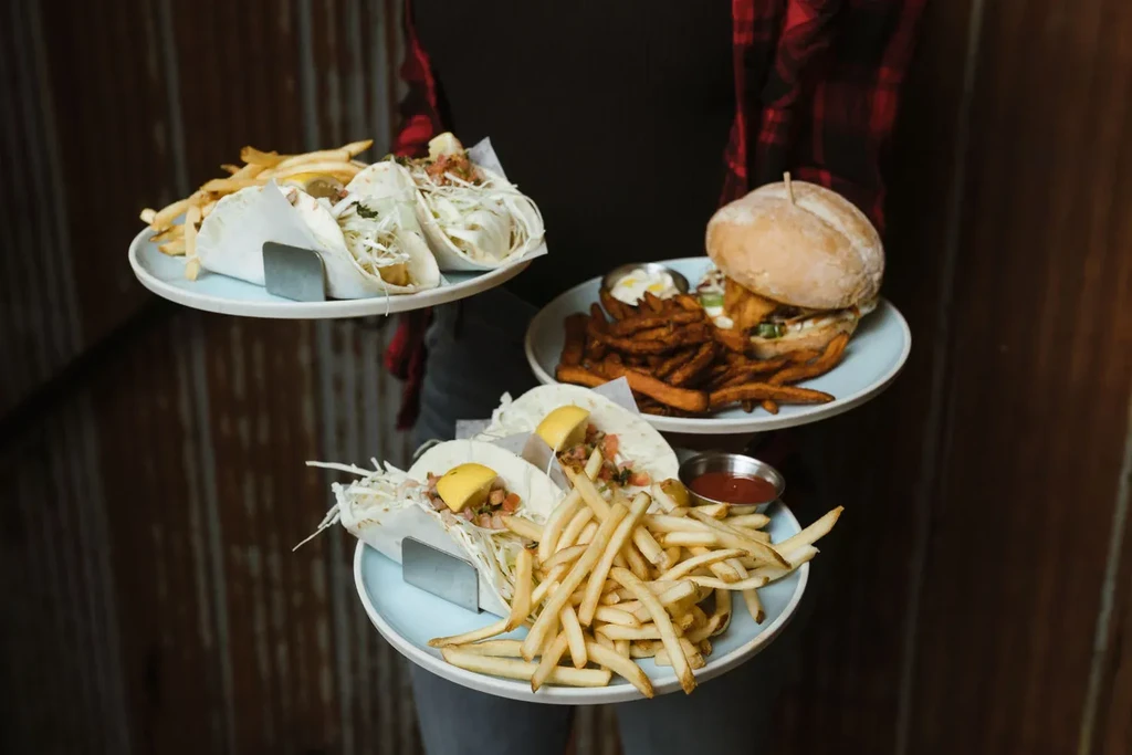 Hands holding three plates: fish tacos, a burger, and fries.