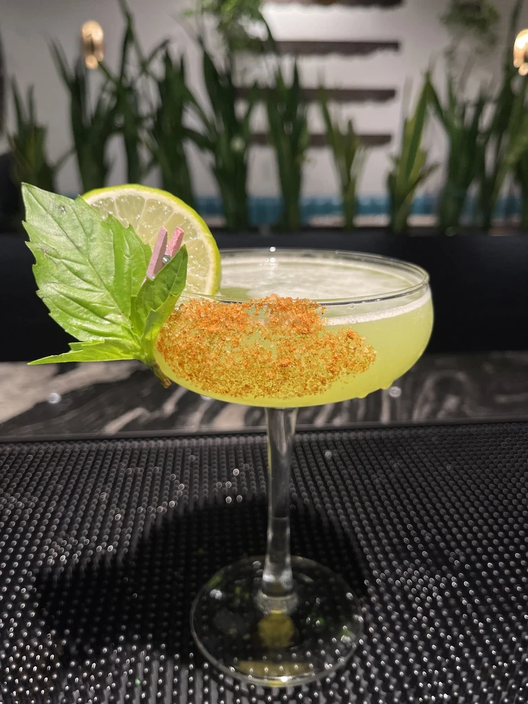 Coupe glass cocktail with granular rim, lime, and pinned leaf.