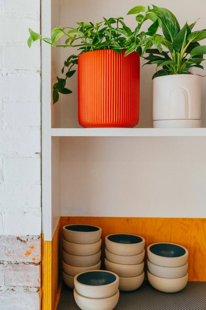 White shelves with potted plants, stacked ceramic bowls, and yellow background.