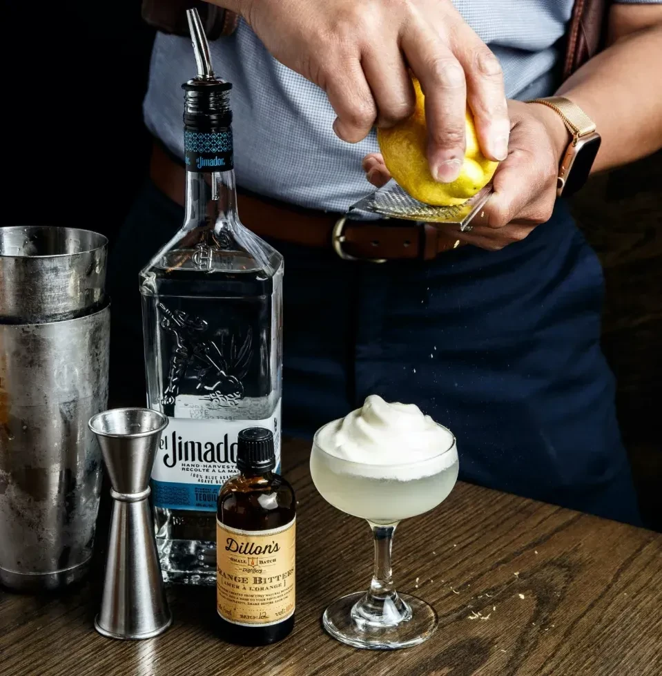 Bartender zesting a lemon over a foamy cocktail with tequila.