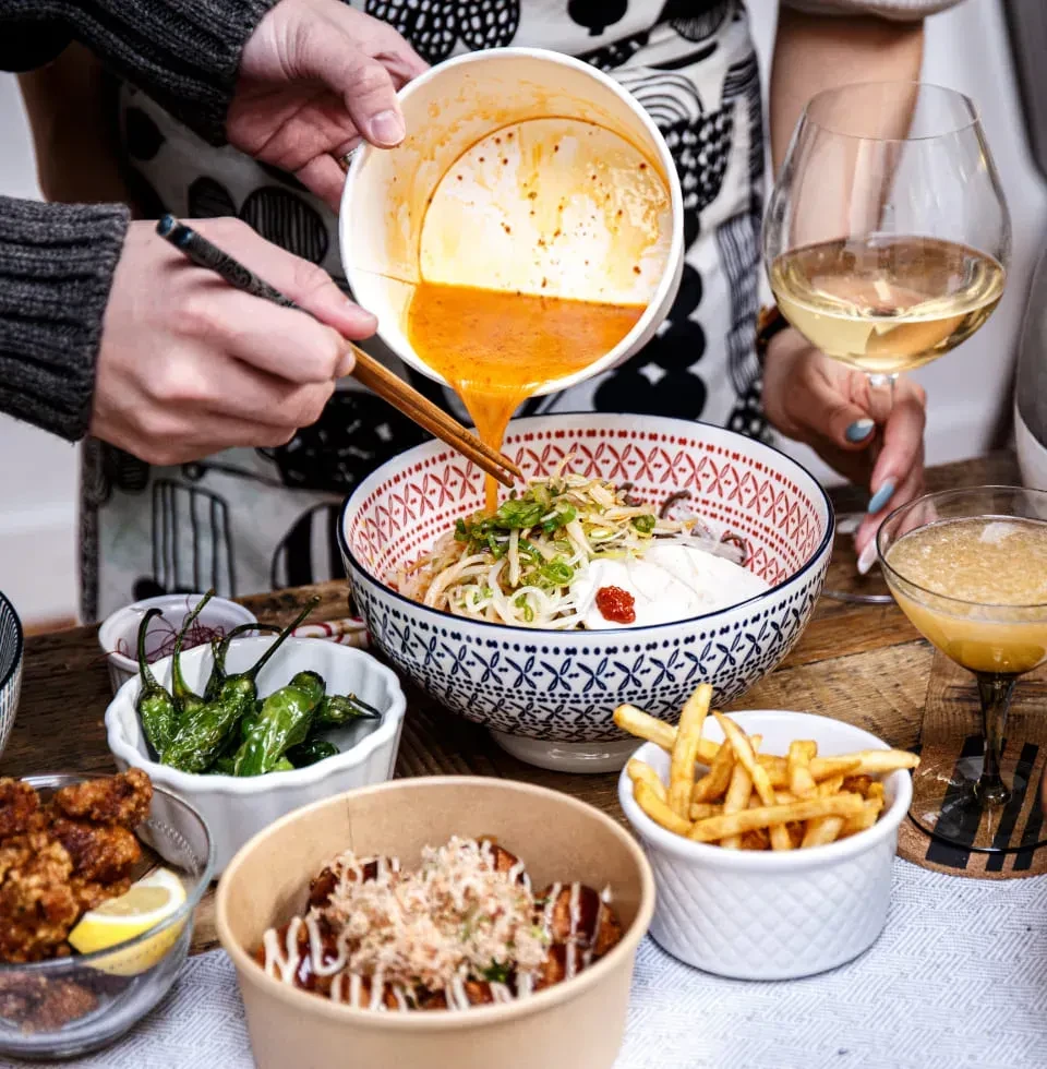 Close-up of hands pouring broth into ramen with other Asian food.