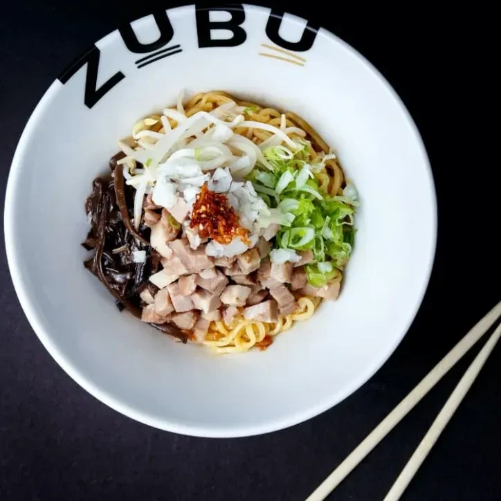 Overhead ZUBU ramen bowl with noodles, pork, vegetables, chili, and chopsticks.