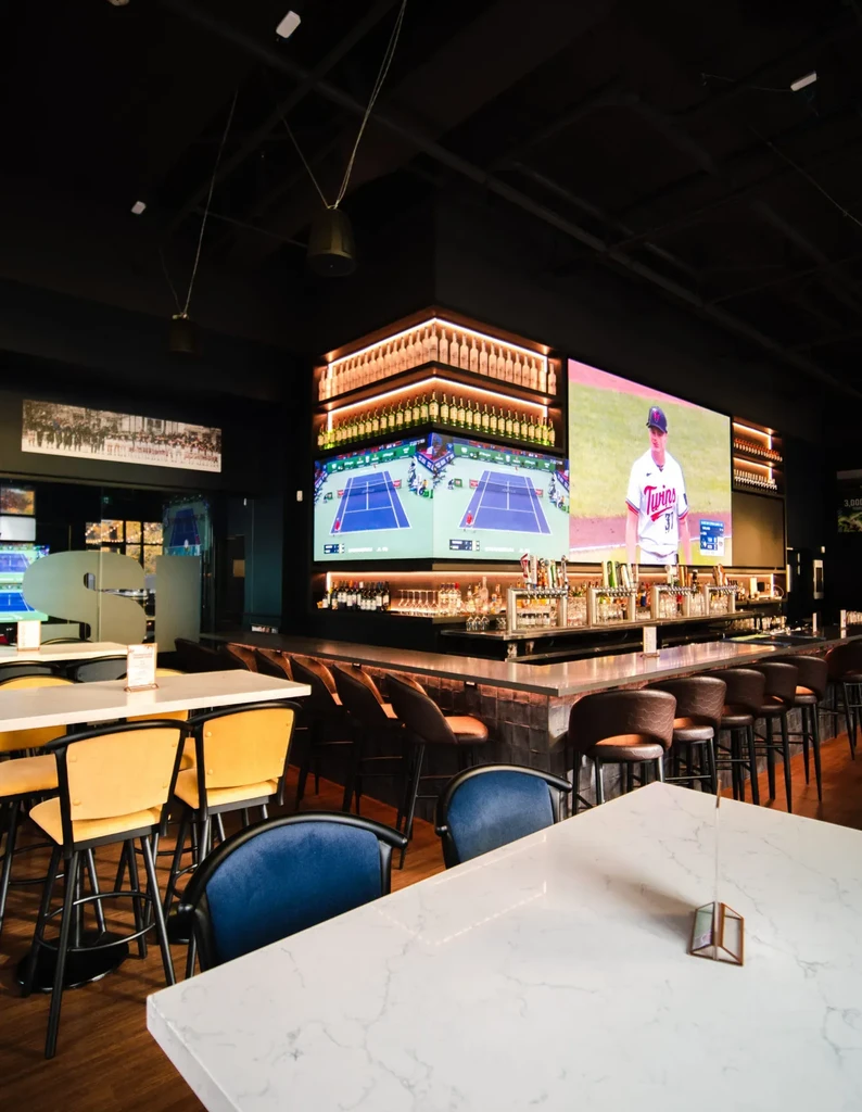 Empty modern sports bar with large screens, backlit bar, and varied seating.