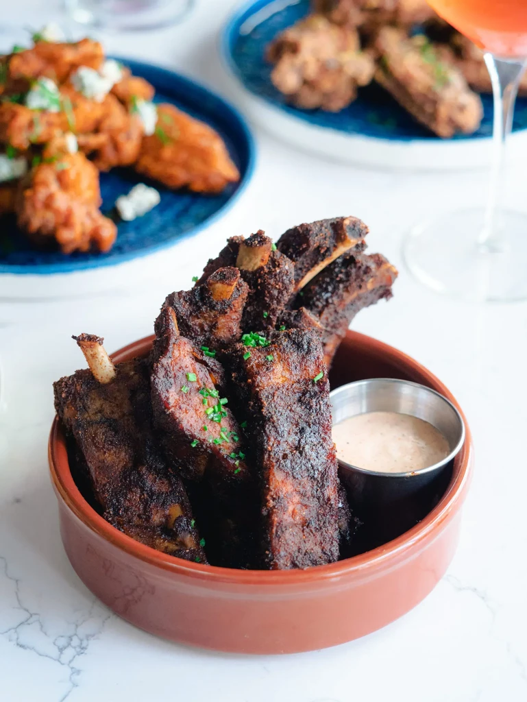 Seasoned ribs, buffalo wings, chicken, and cocktail on a bright tabletop.