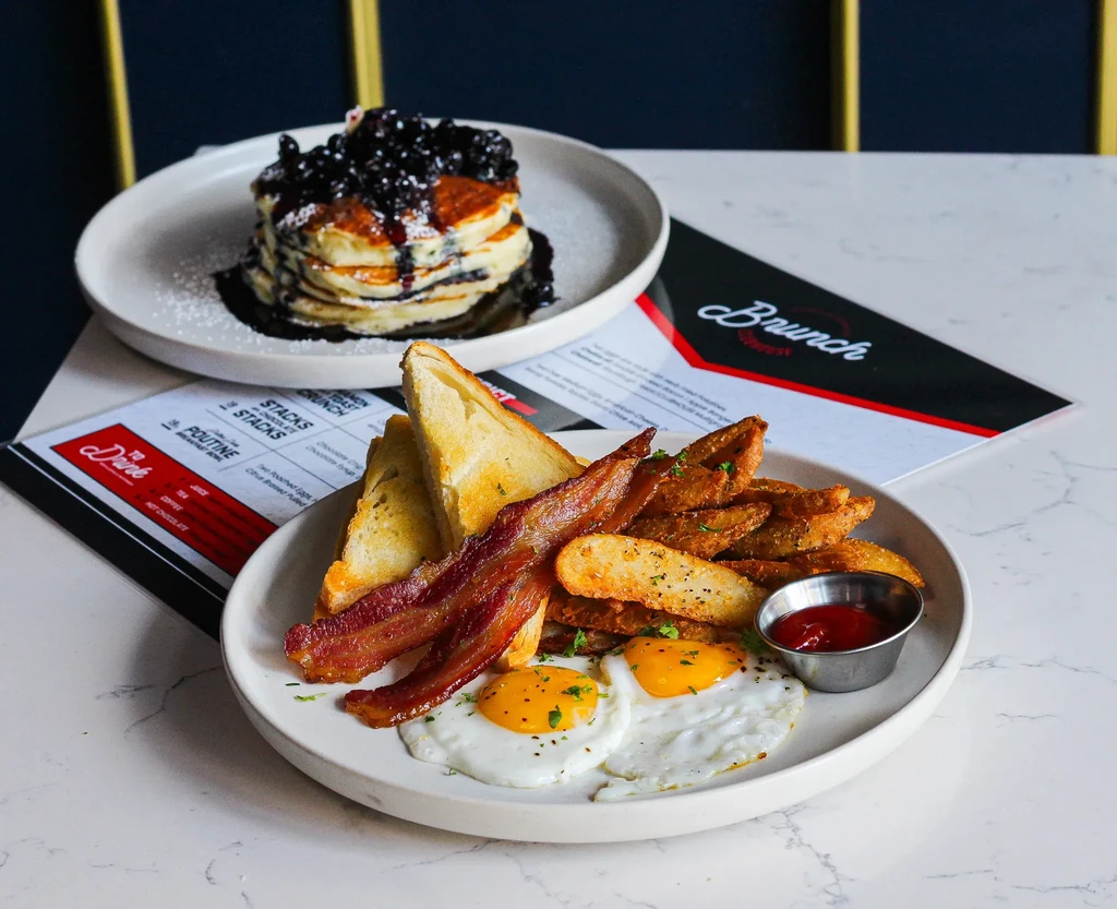 Two plates of brunch food: eggs, bacon, potatoes, and blueberry pancakes.