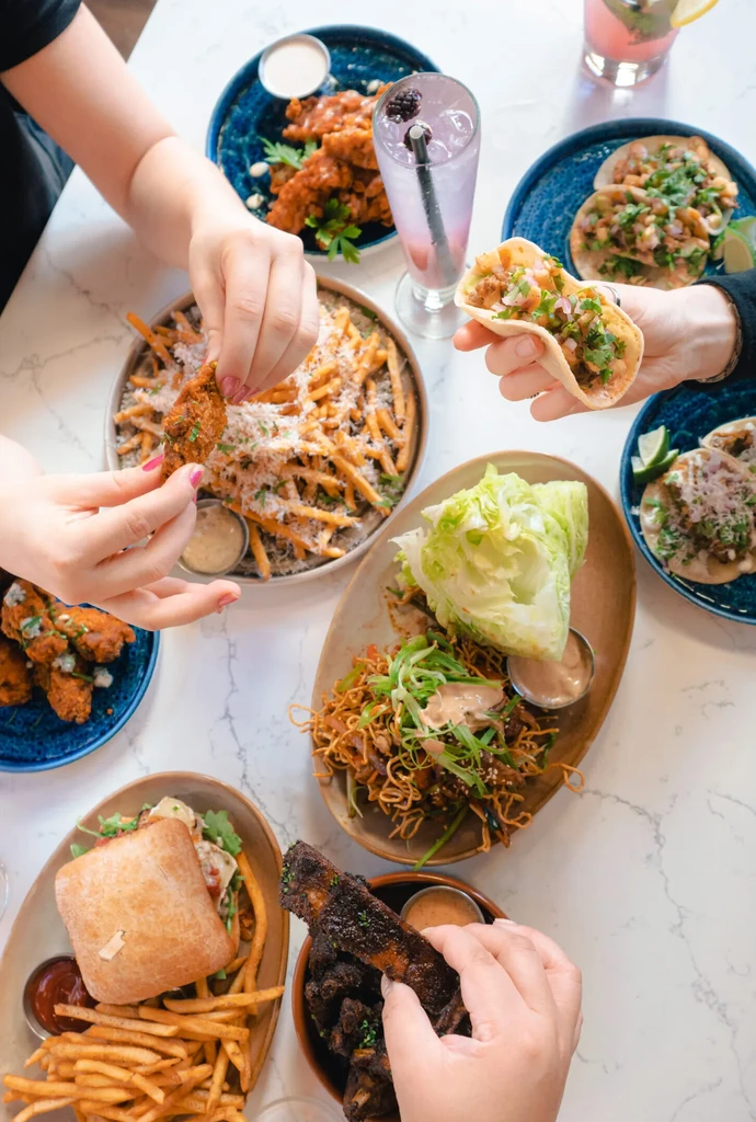 Hands share fried chicken, tacos, fries, and ribs on a marble table.