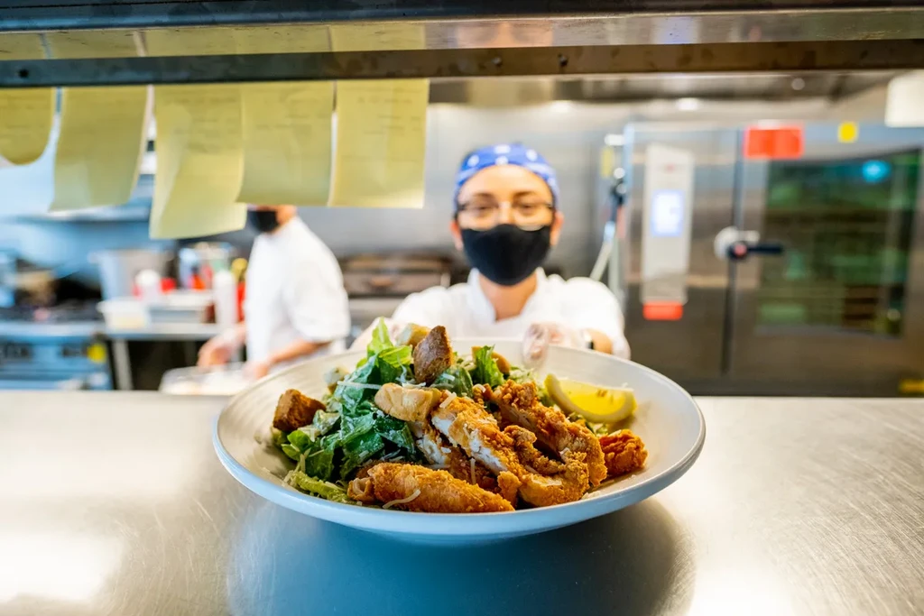 Chef presents Caesar salad with fried chicken and lemon in a kitchen.