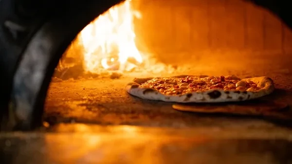 Close-up: Pizza baking in a wood-fired oven, illuminated by bright flames.