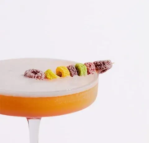 Amber cocktail with white foam, garnished with colorful cereal rings.