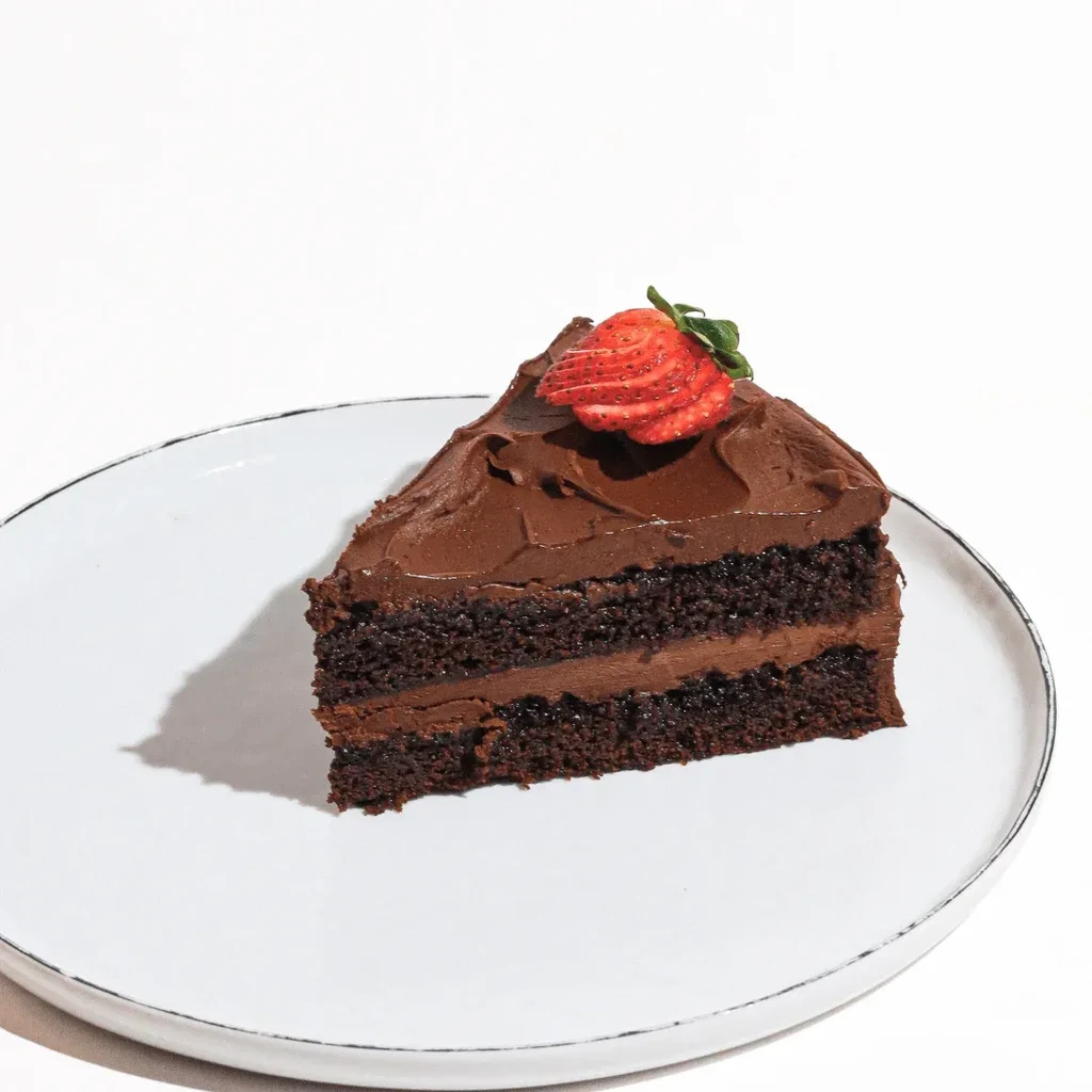 Slice of two-layer chocolate cake with creamy frosting, strawberry, and leaf.
