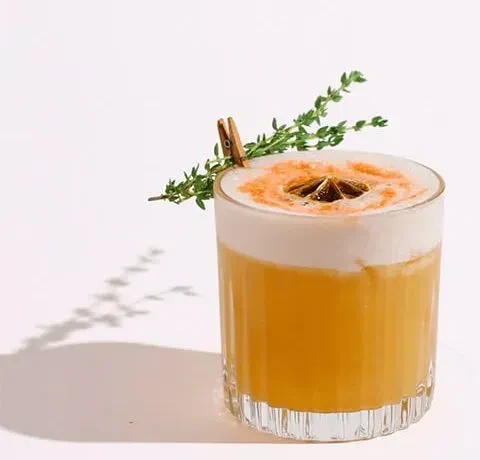 Layered orange-yellow cocktail with white foam, star anise, and herb.