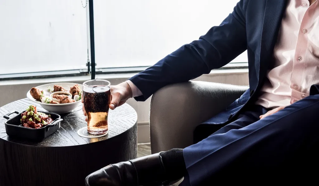 Man in suit holds a bubbly drink, fried chicken, and stew.