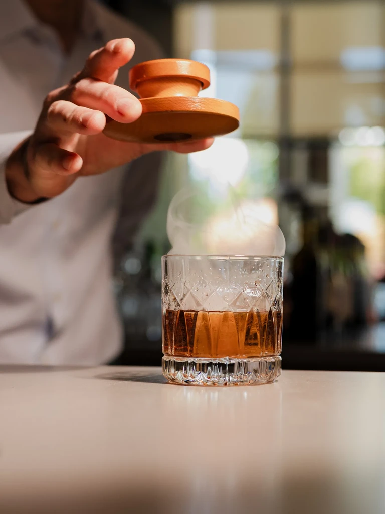 Hand releasing smoke from wooden cloche over amber cocktail.