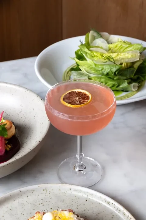 Pink cocktail with dried orange slice and dishes on a table.