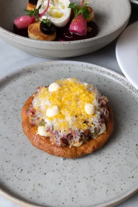 Puffy base topped with tartare, grated cheese, yellow garnish, white sauce.