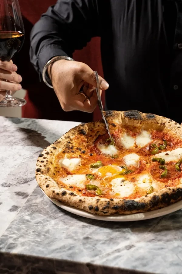 Person cutting Neapolitan pizza with egg yolk, green peppers on marble.