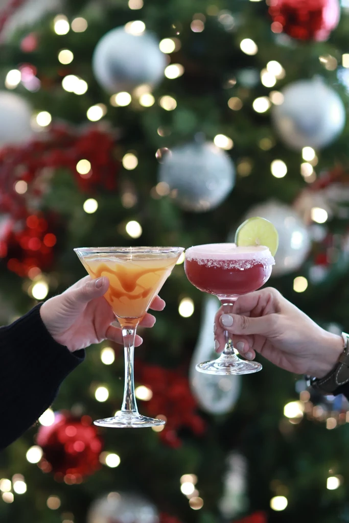 Hands toasting a yellow martini and red coupe cocktail by a Christmas tree.