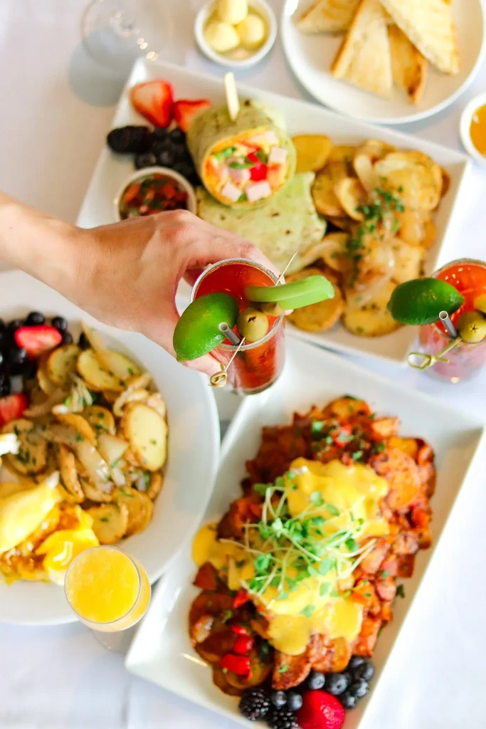 Overhead view of diverse brunch spread; hand holds a Bloody Mary.