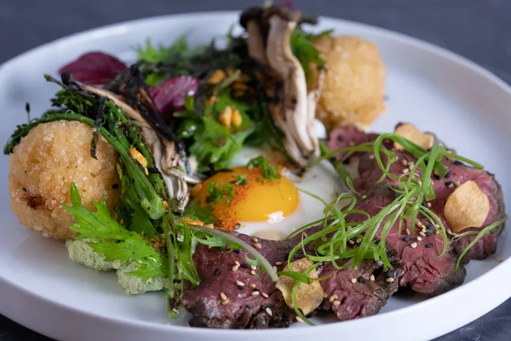 Colorful plated dish with seared meat, egg, fried items, and vegetables.