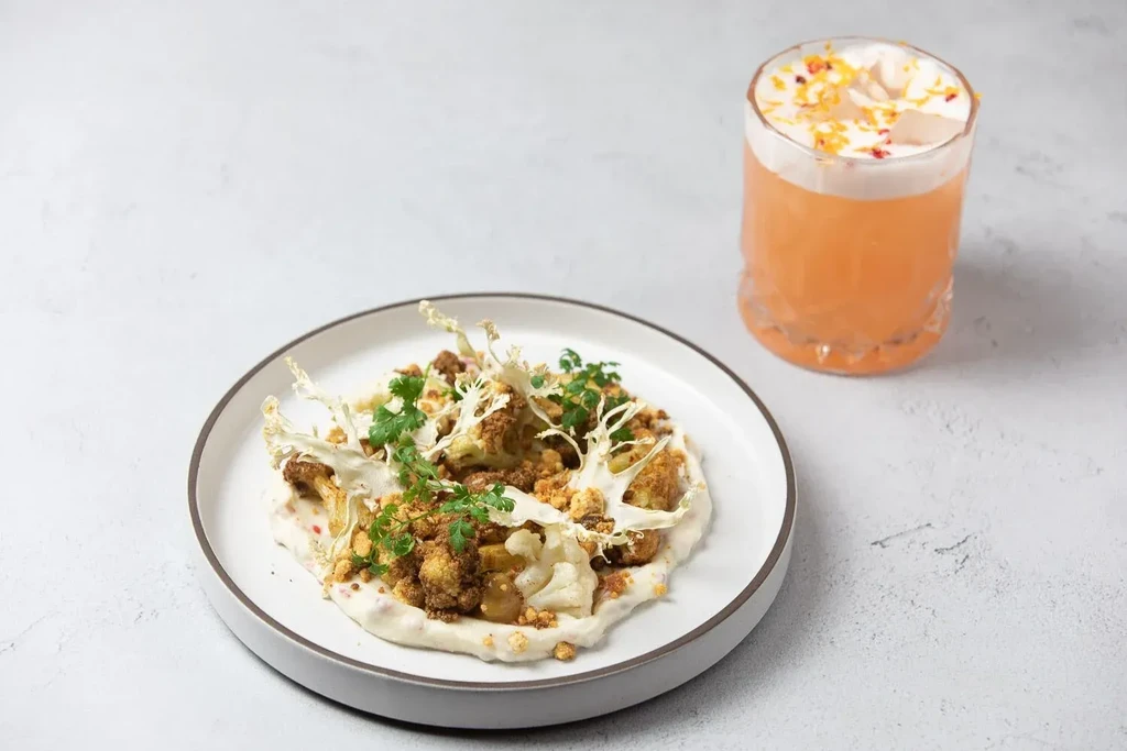 A plated dish of roasted cauliflower and a foamy orange cocktail.