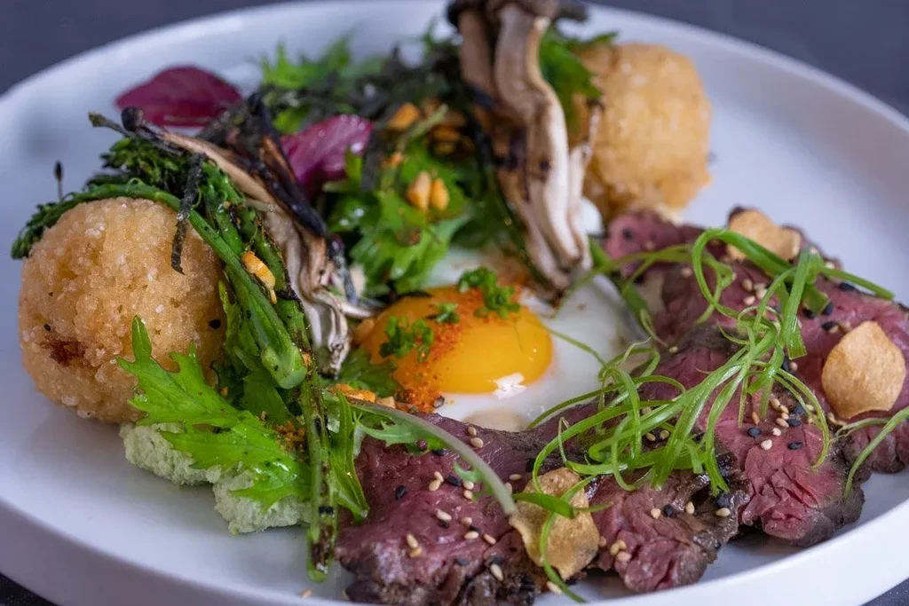 Artfully plated gourmet dish with sliced beef, egg, mushrooms, and greens.