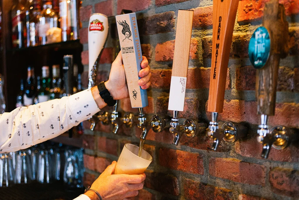 Hands pouring amber beer from a Talisman tap into a frosted glass.
