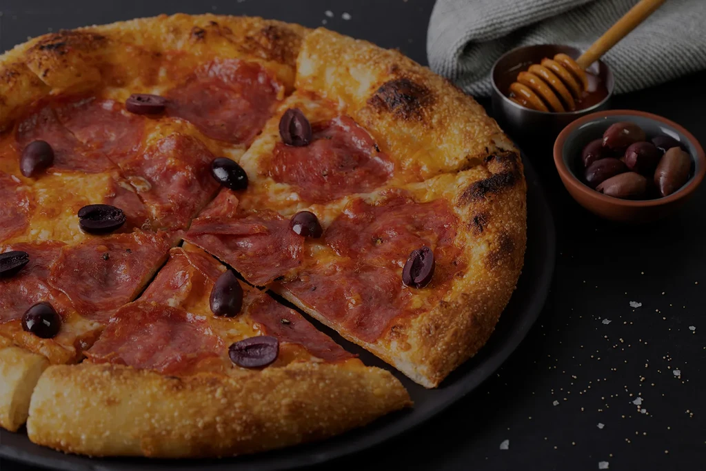 Freshly baked pepperoni and olive pizza with honey dipper and olives.