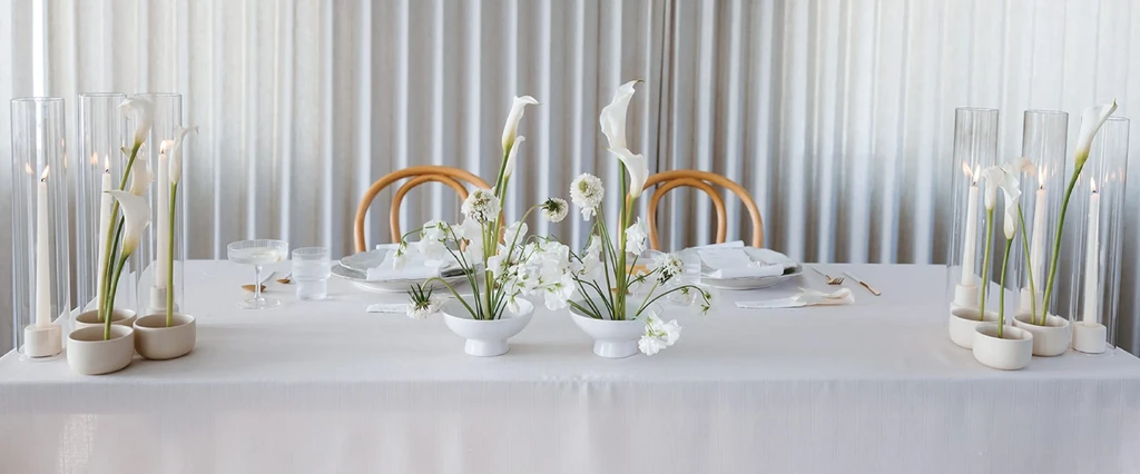 White-clothed dining table with white flowers, candles, and place settings.