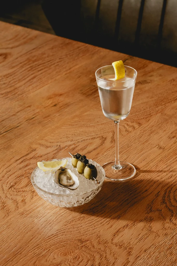 Cocktail, oyster, and caviar-topped olives on a wooden table.