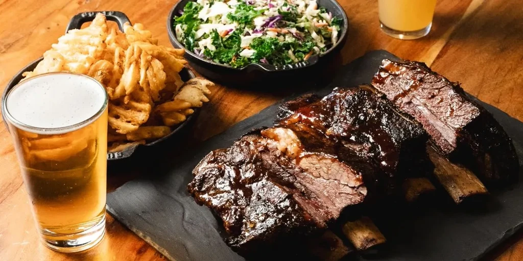 Glazed short ribs, waffle fries, coleslaw, and beer on a rustic table.