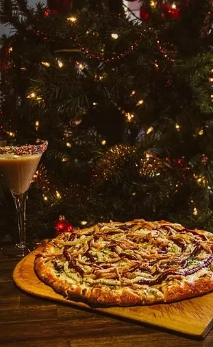 Close-up of a pizza, sprinkle-rimmed drink, and blurred Christmas tree.
