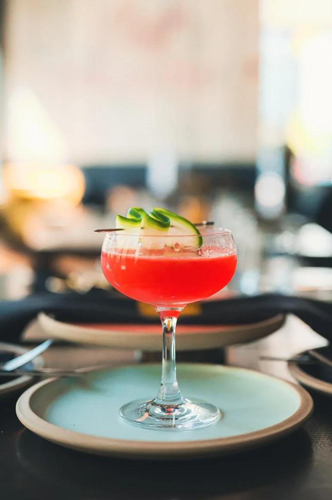 Red cocktail with cucumber garnish on a plate in a bar setting.