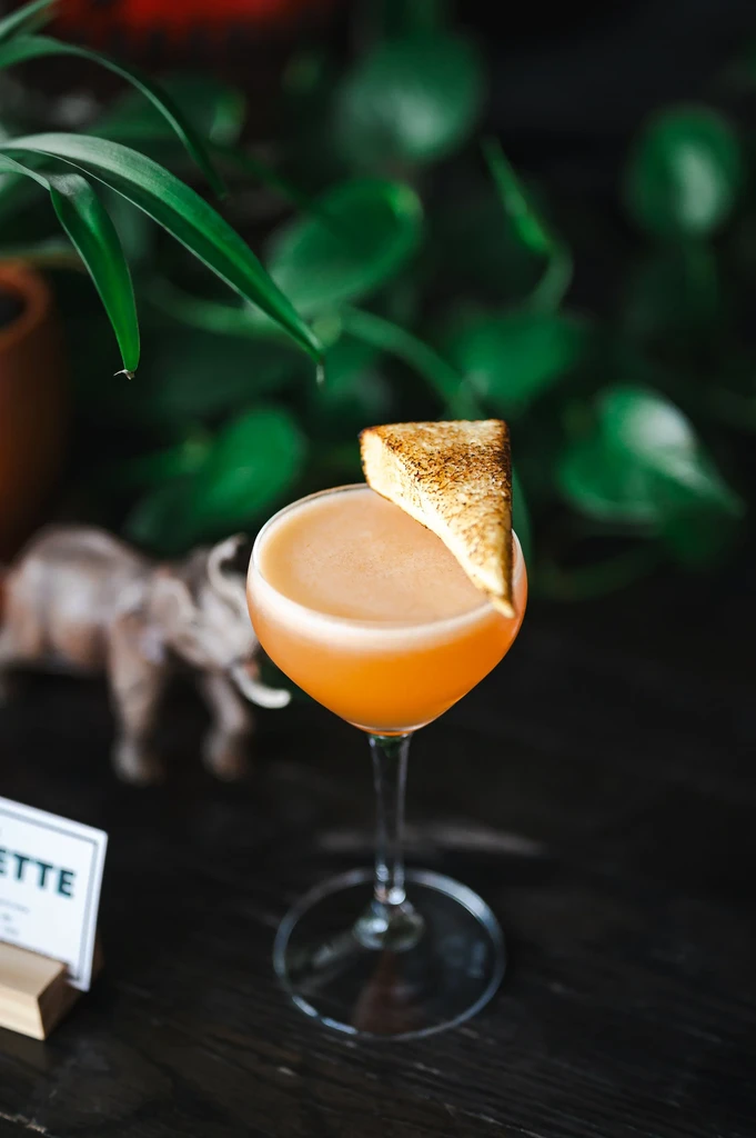 Frothy orange cocktail with toast garnish and blurred green foliage.