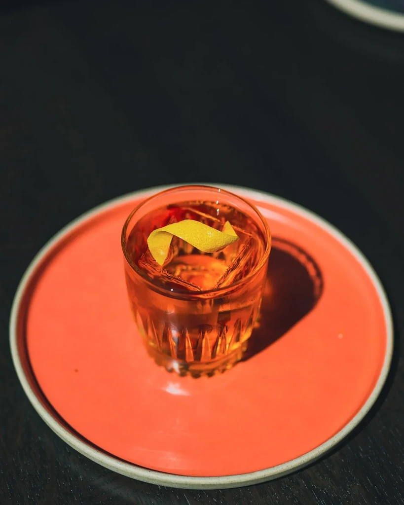 Faceted glass amber cocktail with lemon, ice, on vibrant coral plate.