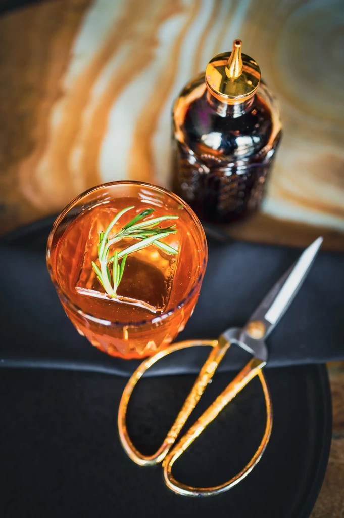 Amber cocktail with square ice, rosemary, bitters bottle, and golden scissors.