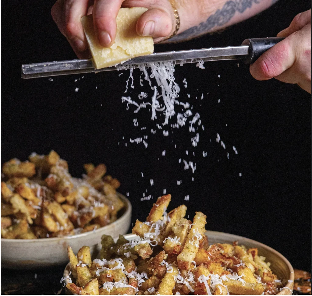 Hands grating light yellow cheese onto golden french fries.