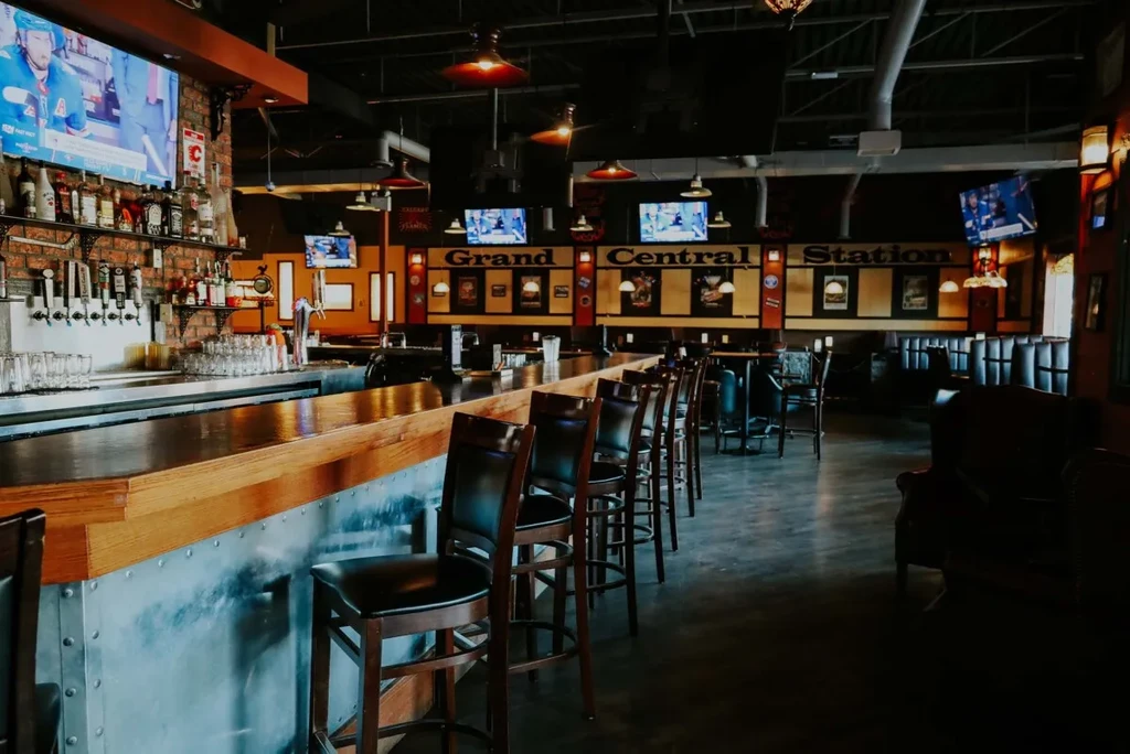 Industrial bar interior: wooden counter, riveted metal, liquor, TVs showing hockey.