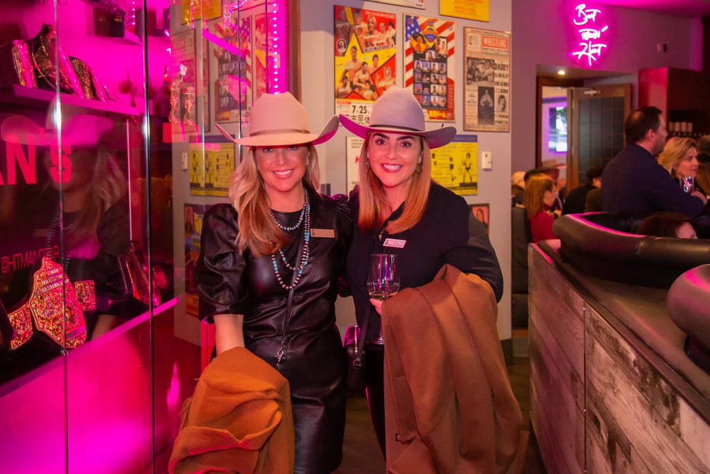 Two smiling women in cowboy hats in a boxing-themed neon bar.