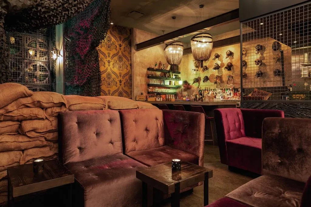 Bar interior with sandbag seating, camouflage, gas mask decor, and dim lighting.