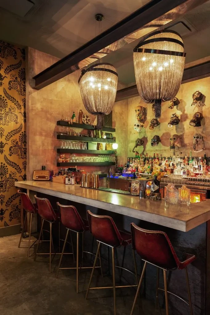 Eclectic bar with concrete counter, damask wallpaper, and steampunk gas masks.