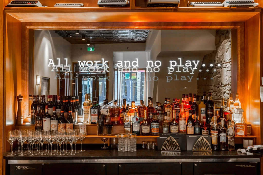 Well-lit, stocked bar with spirits, empty glasses, antique typewriters, and text.