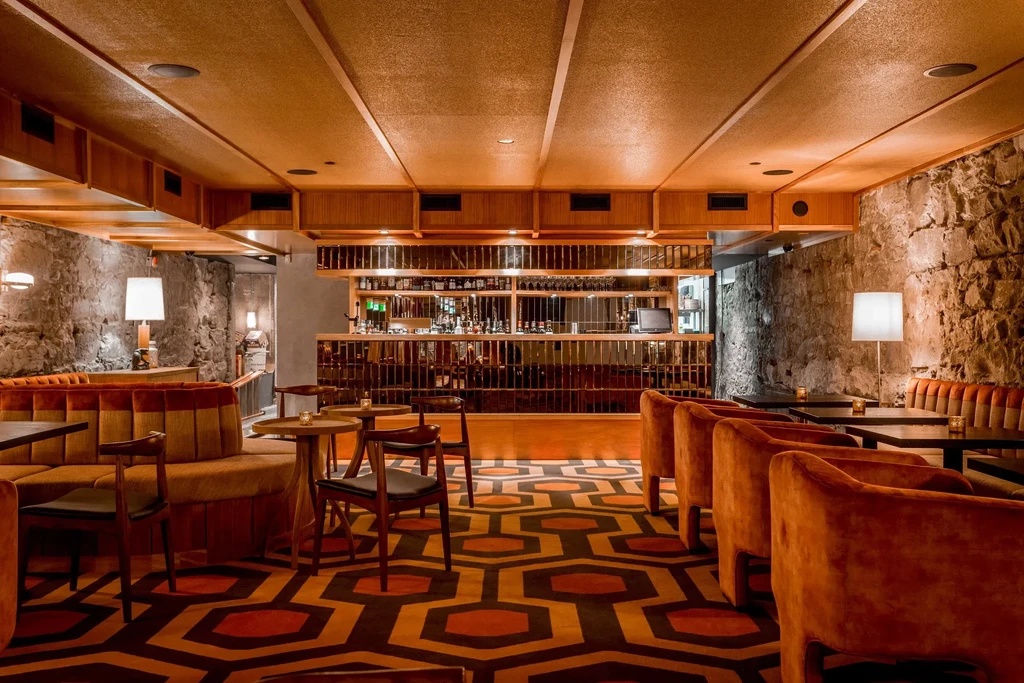Retro bar with geometric carpet, orange velvet seating, and stone walls.