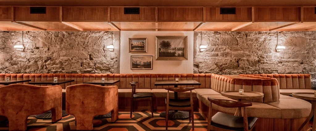 Empty restaurant interior with tufted reddish-brown banquette, stone wall, hexagonal floor.