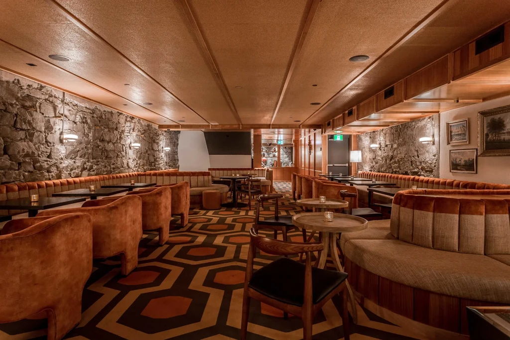 Bar lounge with stone walls, wood ceilings, velvet chairs, and patterned carpet.