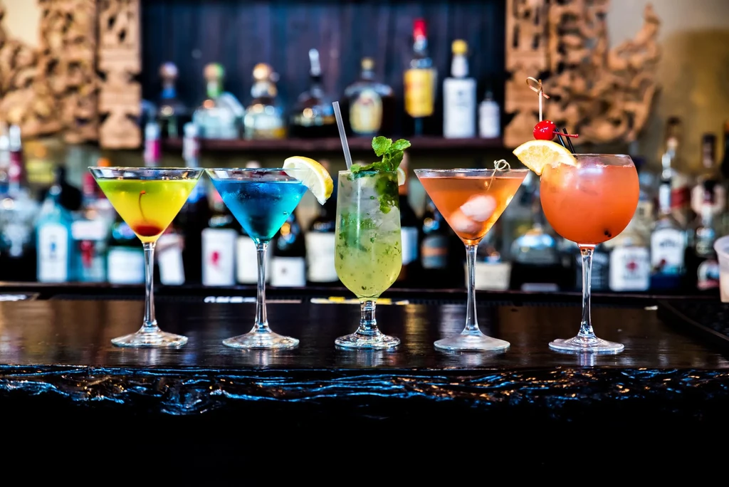 Five vibrantly colored cocktails lined up on a dark bar counter.
