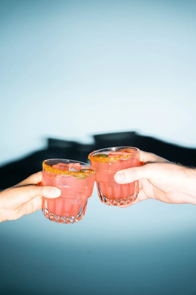 Hands clinking two pink cocktails garnished with spiced rims and jalapeños.
