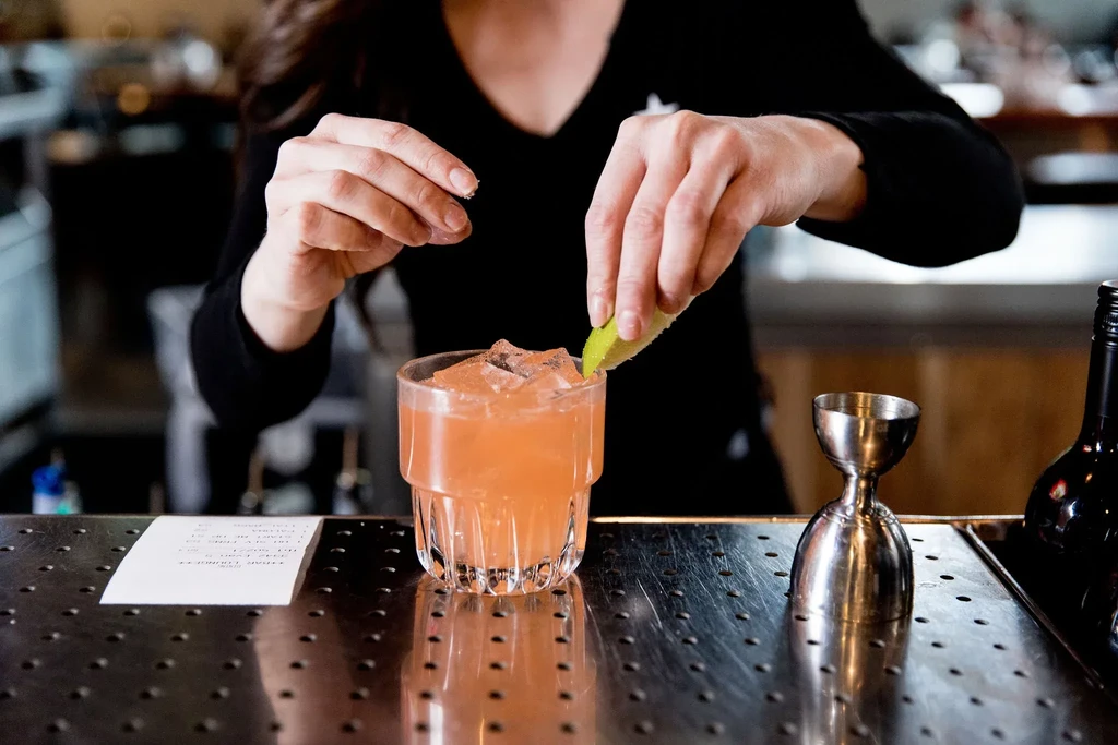 Bartender's hands garnishing a coral-orange cocktail with lime and ice.