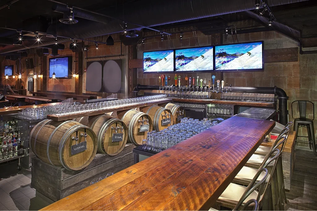 Modern rustic bar counter with beer taps, wine barrels, and TVs.
